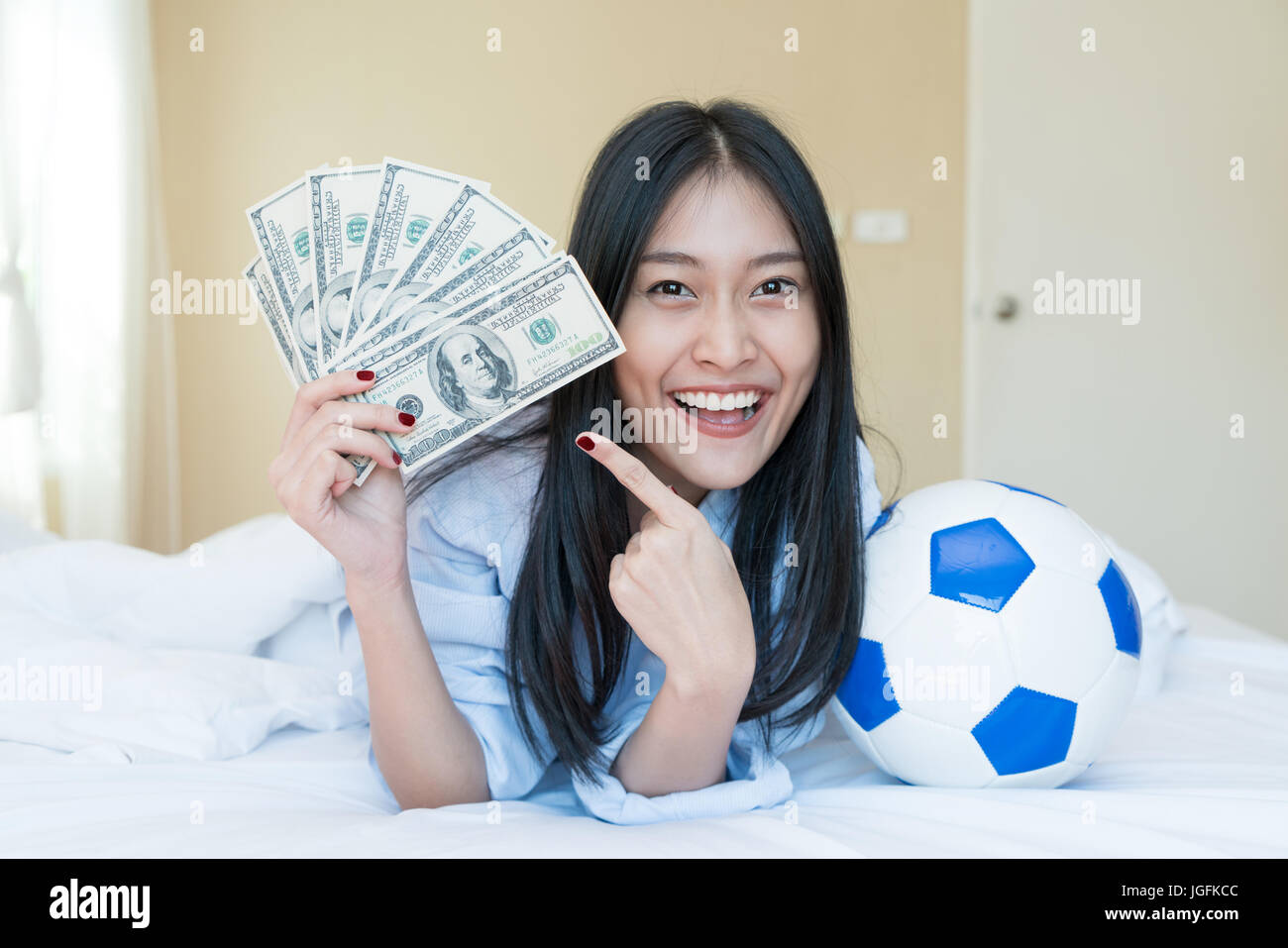 Football and money. Young Asian woman holding soccer ball and cash in bedroom at home. Competition lottery gambling concept idea Stock Photo