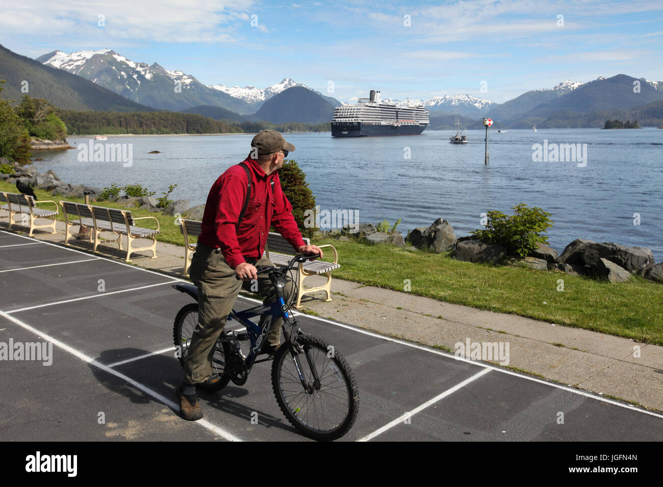 A man on a bicycle looks at a cruise ship anchored in Sitka's harbor in front of snow-covered mountains. Stock Photo