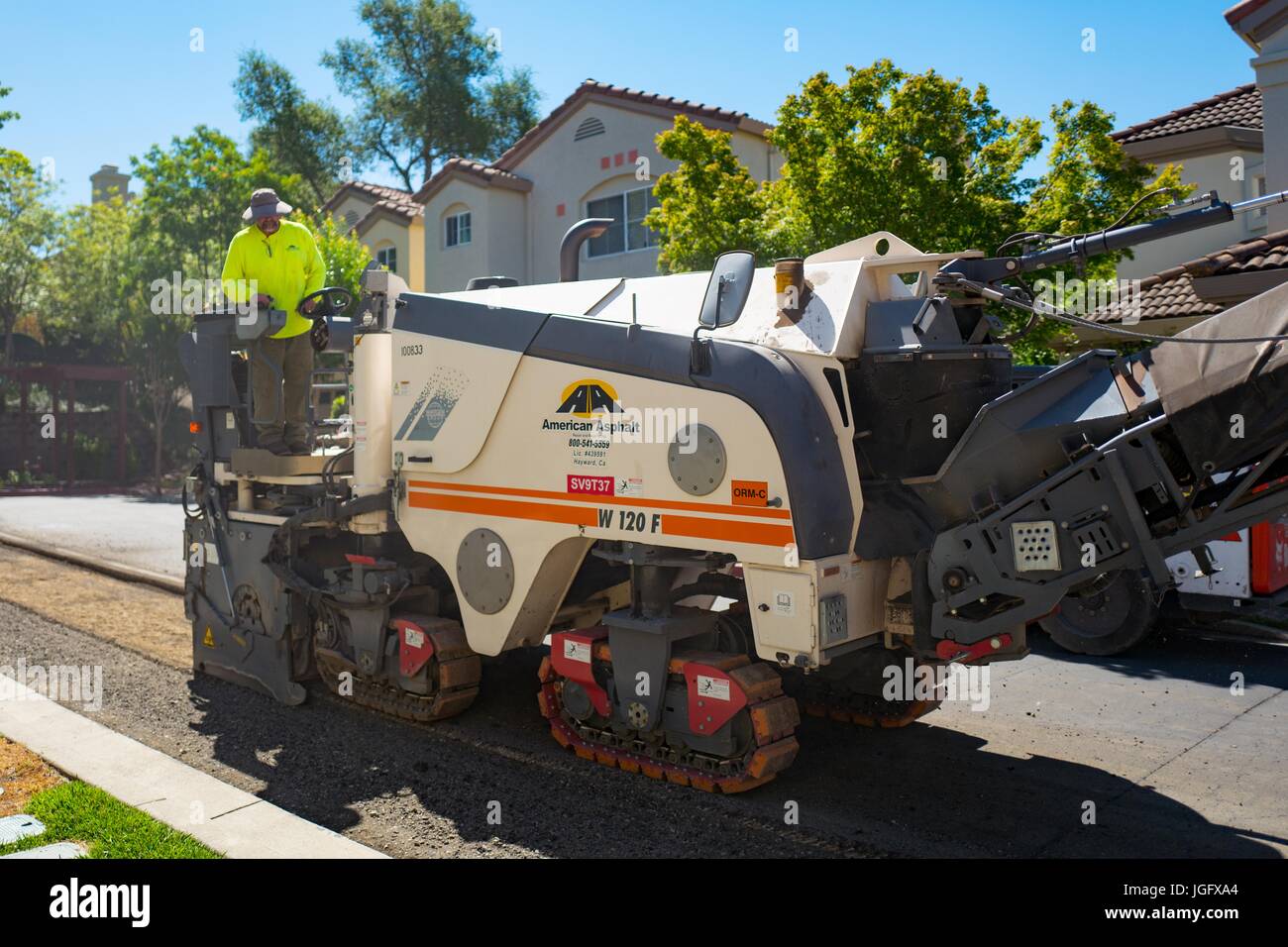 A construction worker for American Asphalt, wearing a bright yellow high visibility vest, steers a machine as it removes asphalt during a road construction and resurfacing project in the San Francisco Bay Area suburb of San Ramon, California, June 26, 2017. Stock Photo
