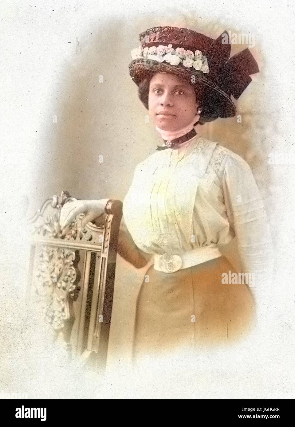 Half length standing portrait of young African American woman next to chair, wearing a blouse, a skirt, an elaborate hat, earrings, and a neutral expression, possibly Philadelphia, Pennsylvania, 1915. Note: Image has been digitally colorized using a modern process. Colors may not be period-accurate. Stock Photo