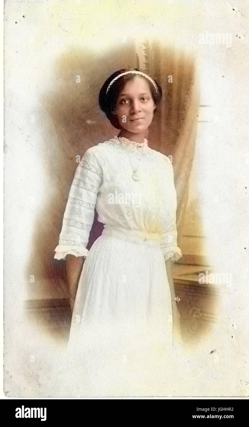 Young African-American woman in white dress, hands behind her back, with a serious expression, wearing a white headband, 1915. Note: Image has been digitally colorized using a modern process. Colors may not be period-accurate. Stock Photo
