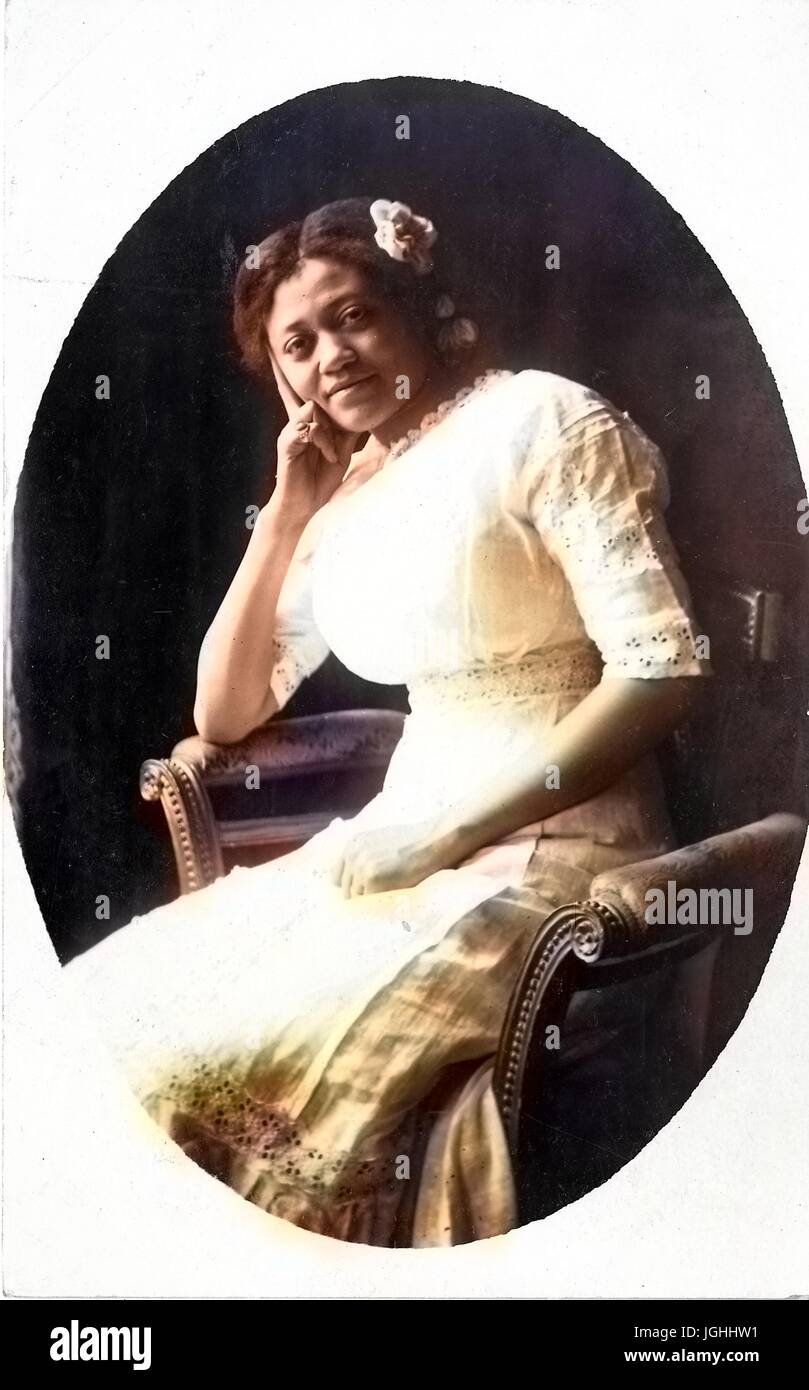 Three quarter seated portrait of smiling young African American woman, wearing a light dress and a flower in her hair, with her head tilted onto her right hand, seated in patterned chair, 1915. Note: Image has been digitally colorized using a modern process. Colors may not be period-accurate. Stock Photo