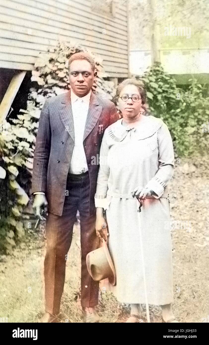 African American husband and wife standing next to each other in front of a wood building and shrubbery, the woman is wearing a light-colored shirt and long skirt and is holding a hat in one hand and a cane in the other, the man is wearing a full dark-colored suit with a light colored shirt underneath and he is holding a cigar in one hand, 1950. Note: Image has been digitally colorized using a modern process. Colors may not be period-accurate. Stock Photo