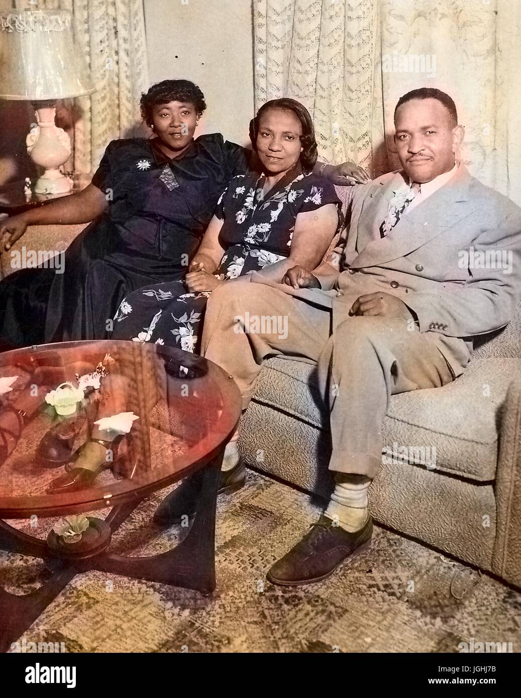 African-American family, father, daughter, and mother sitting on a couch in their home, glass coffee table with figurines visible in the foreground, a lamp and curtains in the background, 1960. Note: Image has been digitally colorized using a modern process. Colors may not be period-accurate. Stock Photo