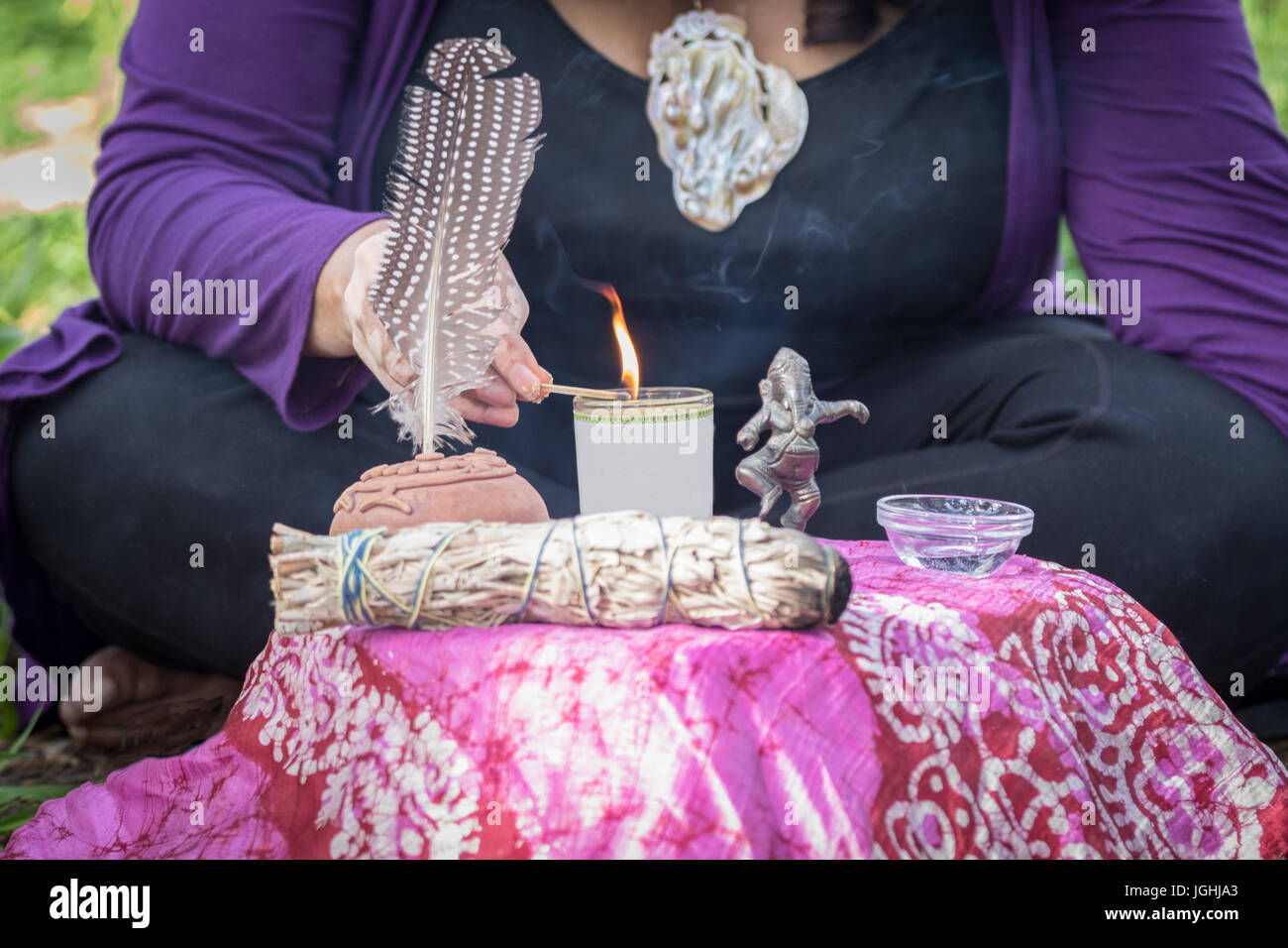 Female shaman lighting candle at altar seated on the ground. You can see the flame the represents fire, along with water, ganesha and other symbols. Stock Photo