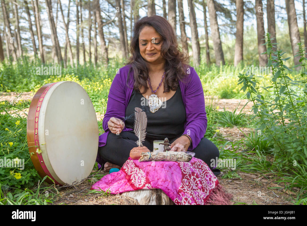 Female shaman smiles as she lights a candle at altar seated on the ground in a forest Stock Photo