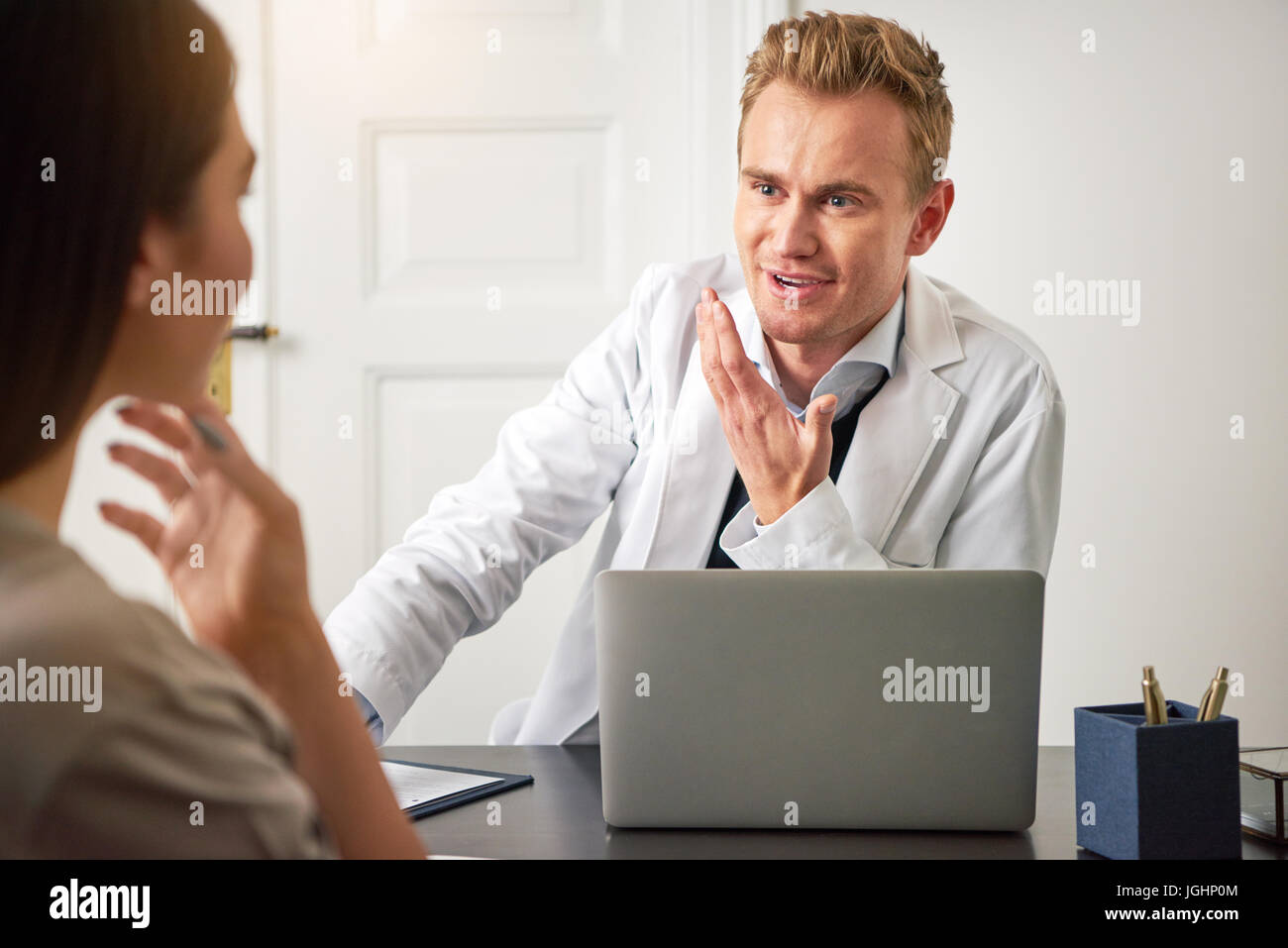 Young smiling cosmetologist man sitting at a laptop in the office and communicating with a woman. Stock Photo