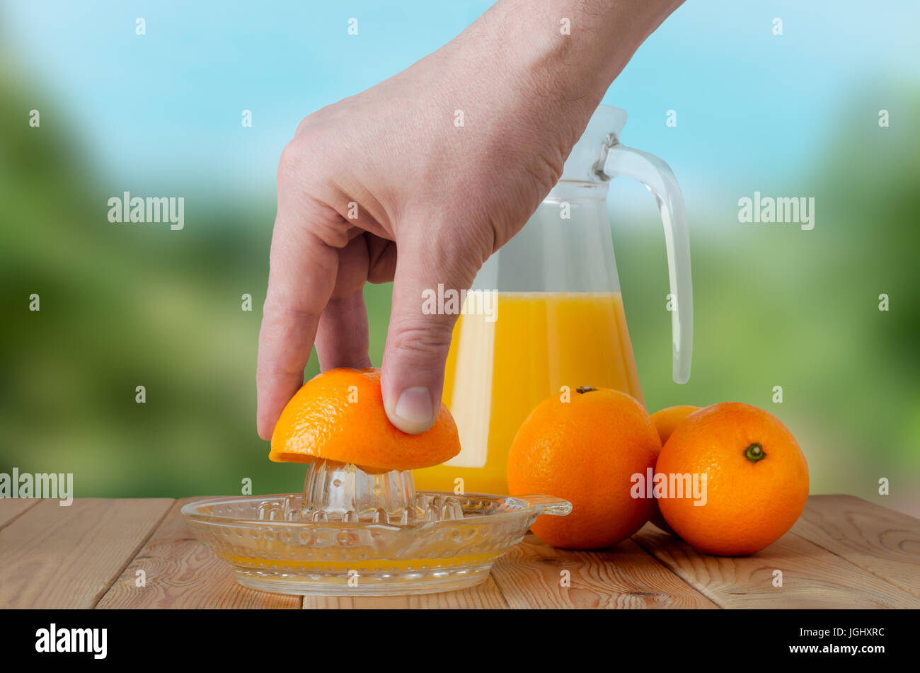 A hand squeezing juice from an orange on a manual glass squeezer.  Set on a wooden planked table with a group of three oranges and a glass jug of juic Stock Photo