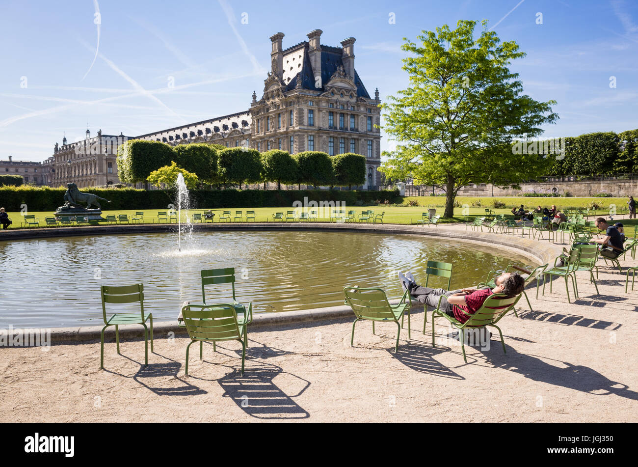 The Tuileries garden in Paris, France, by a sunny morning with a man resting on a metal lawn chair near a water basin and the Louvre palace. Stock Photo