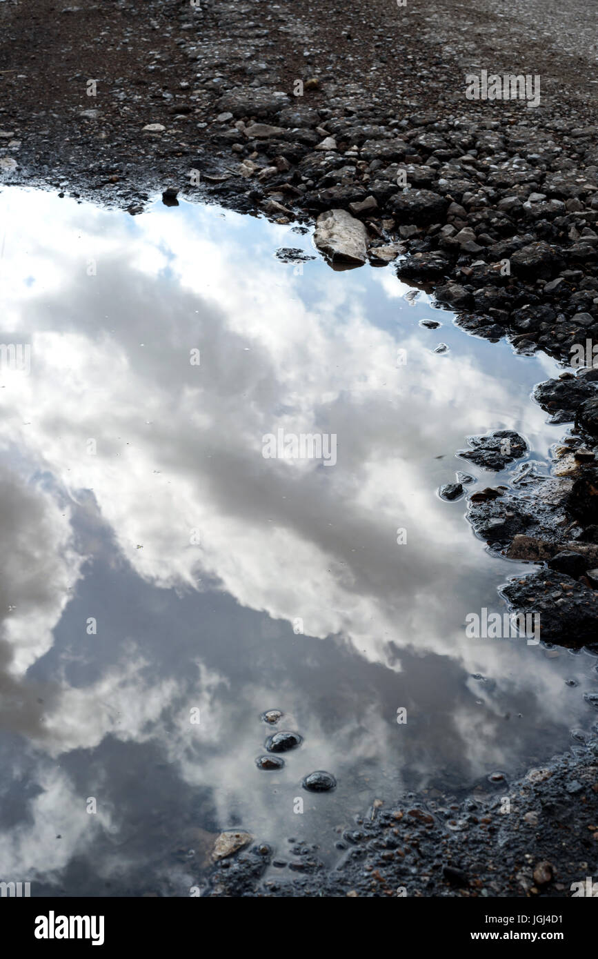 Cracked and broken asphalt pothole on the surface of a crumbling road. Rain water puddle collects in the cracked hole reflecting the sky. Stock Photo