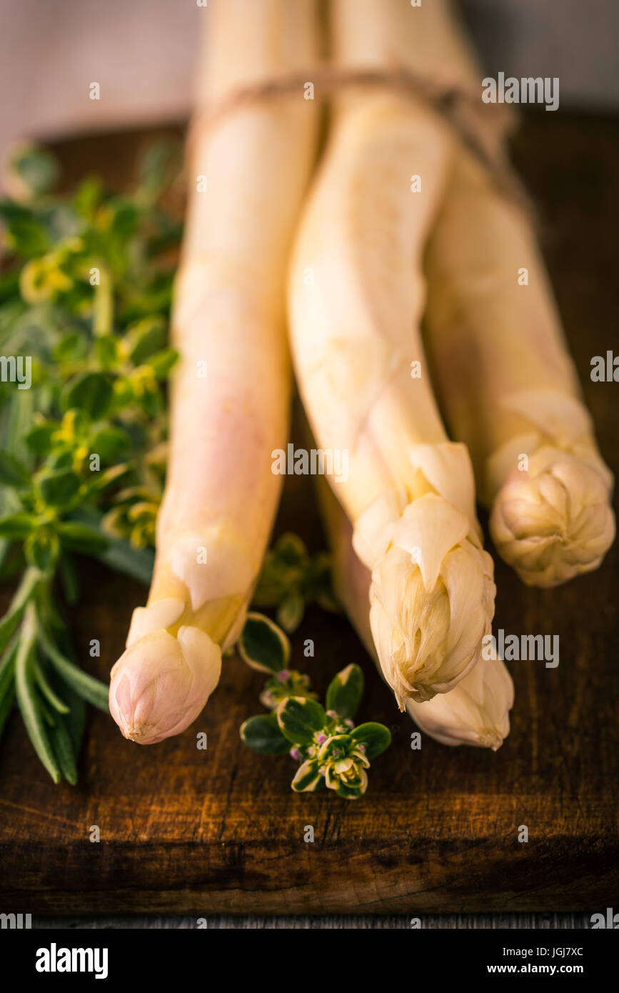 Vertical photo with bonded bunch of white asparagus. Delicious vegetable is placed on dark vintage wooden chopping board with brown color and green he Stock Photo