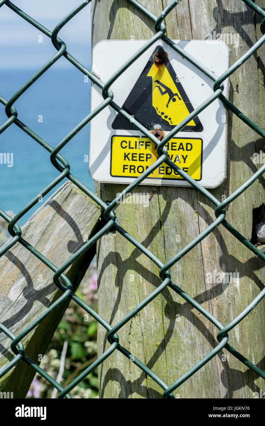 Triangular hazard warning sign behind a mesh fence keeping the public away from cliff edges around Newquay in Cornwall.Metaphor for post-Brexit UK? Stock Photo