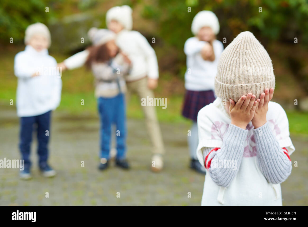 Portrait of little boy crying hiding his face with cruel kids pointing at him in background, bullying and calling him names Stock Photo