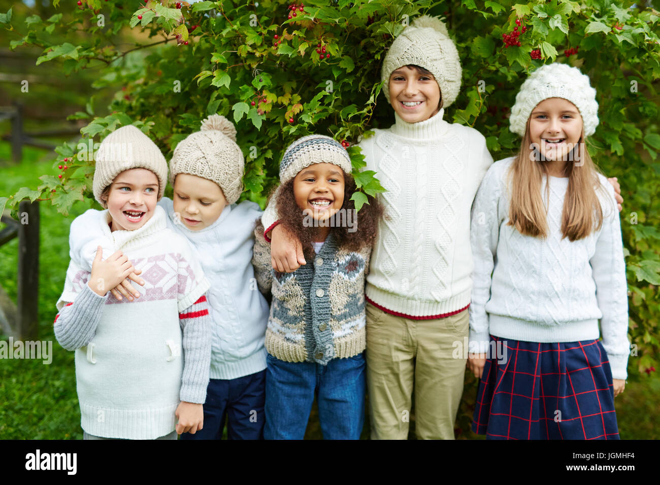 Happy children of various ages posing outdoors embracing and smiling to camera standing under lush green bush enjoying warm autumn day, all dressed in Stock Photo