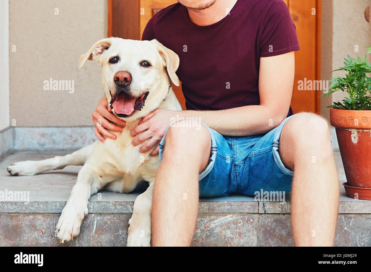 Happy labrador retriever. Young man resting with old dog on the stairs in front of the house. Stock Photo