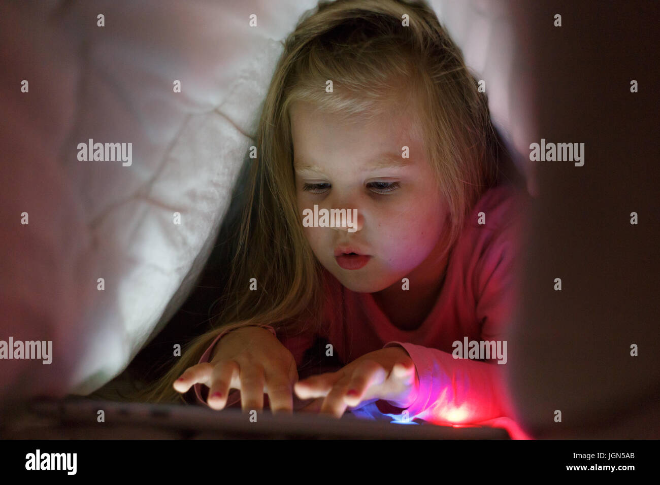 Little girl is playing on the tablet in secret from her parents at night under the blanket. Stock Photo