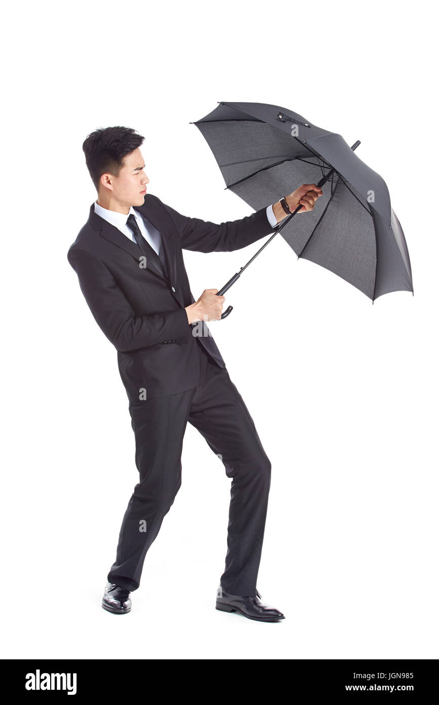 asian business man wearing suit fighting against strong wind holding on to a black umbrella, isolated on white background. Stock Photo