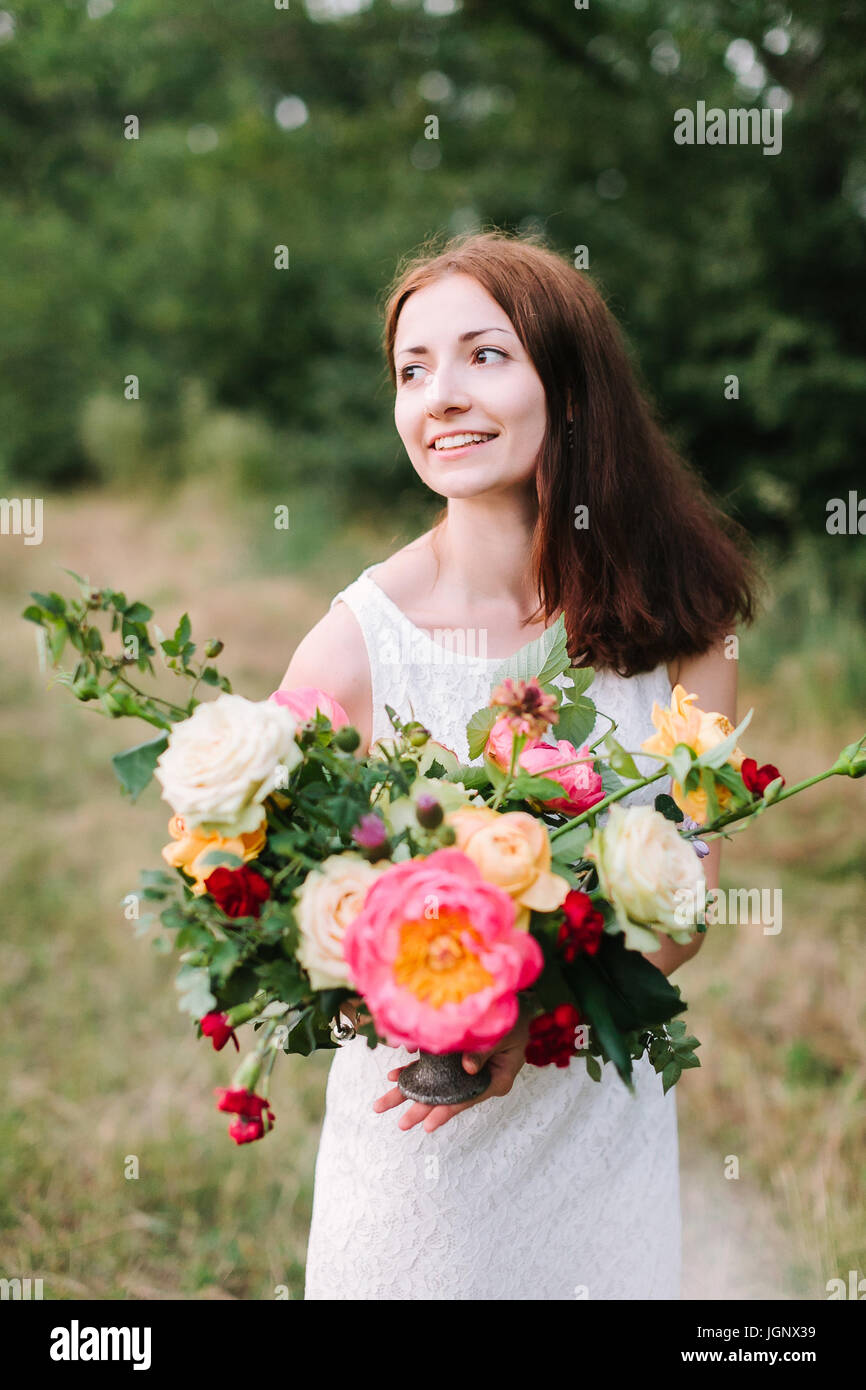 beauty, wedding, spring, nature, decoration concept - shapely smiling woman with dark brown hair in white party dress with colorful bouquet composed o Stock Photo