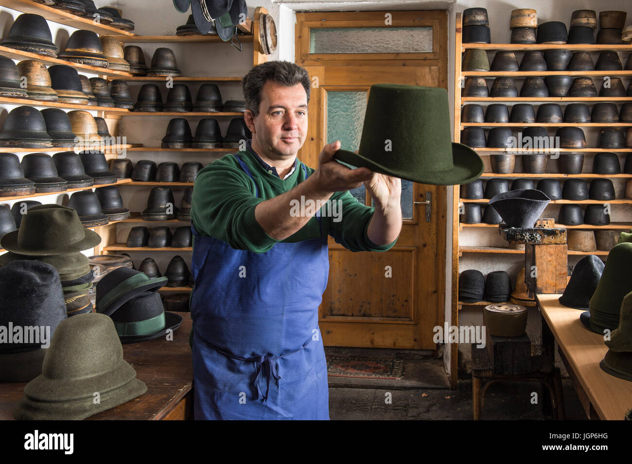 Hatter examining finished wool felt hat, hat molds on wooden shelves and finished hats behind, hatmaker workshop, Bad Aussee Stock Photo