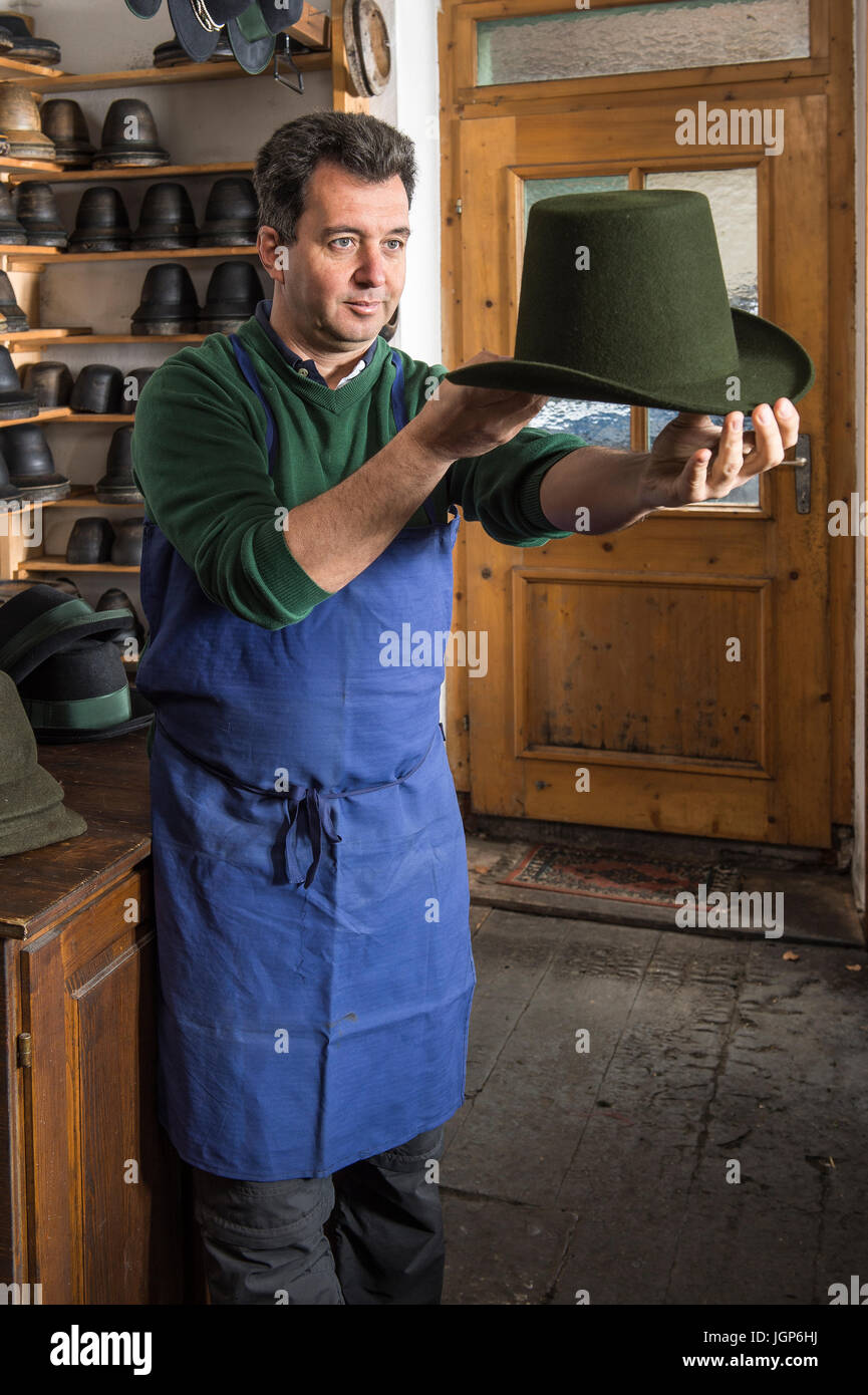 Hatter examining finished wool felt hat, hat molds on wooden shelves and finished hats behind, hatmaker workshop, Bad Aussee Stock Photo