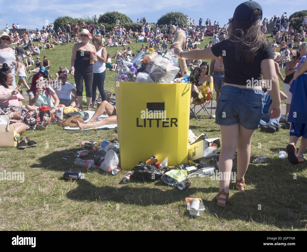A young girl throws rubbish into an overflowing litter bin in a public park  with rubbish lying on the floor next to it. Stock Photo