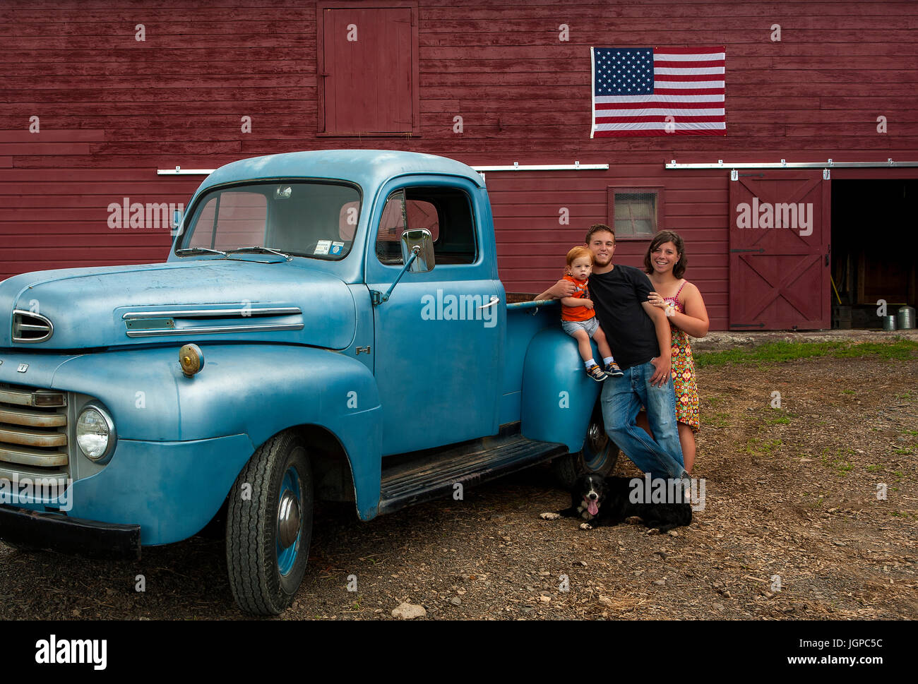 American Farm family with vintage blue truck in front of red barn with American flag, dog at feet, red headed toddler Stock Photo