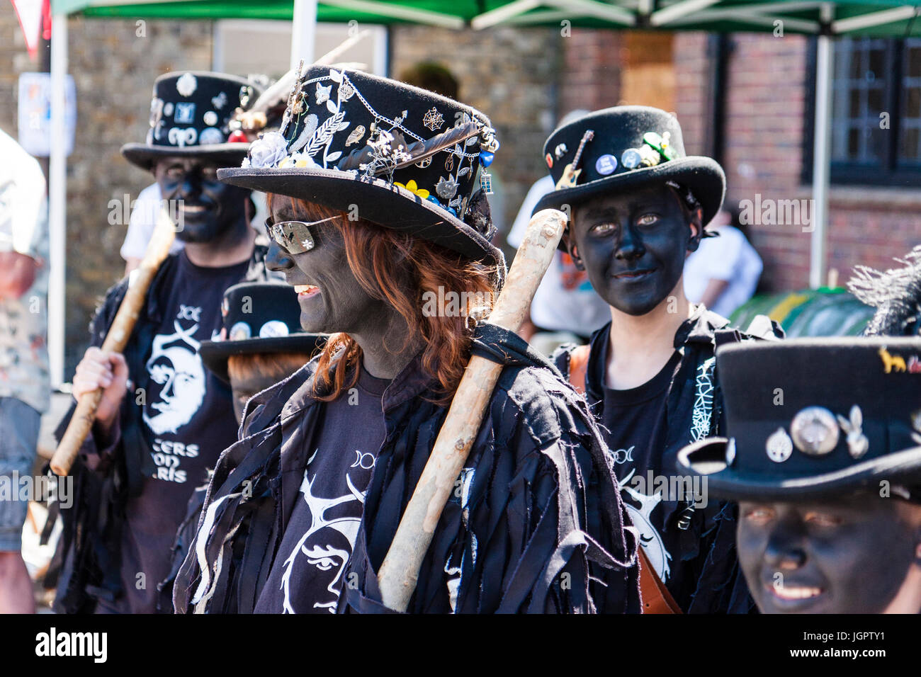 Traditional English folk dancers, Hunters Moon Morris dancers, close up of smiling woman with blacked face, black hat and t-shirt, teenage boy behind her looking at viewer, eye-contact. Stock Photo
