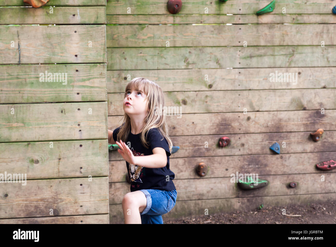 Little girl starting to climb up a wooden climbing wall. Cute child with physical active lifestyle outdoors Stock Photo