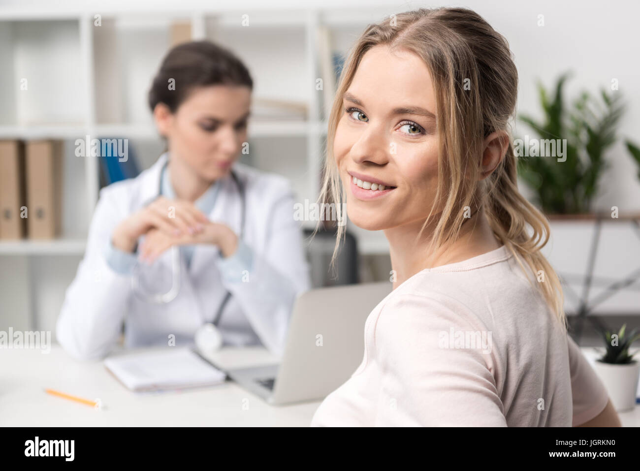 Close-up view of beautiful young patient smiling at camera and doctor using laptop behind Stock Photo