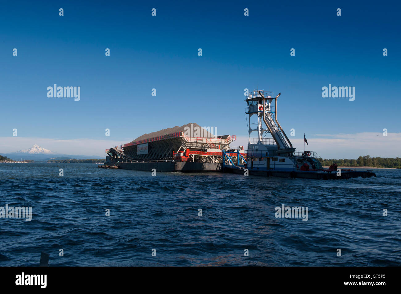 A towboat pushes an aggregate loaded barge up the Columbia River with snow capped Mt. Hood in the distance Stock Photo