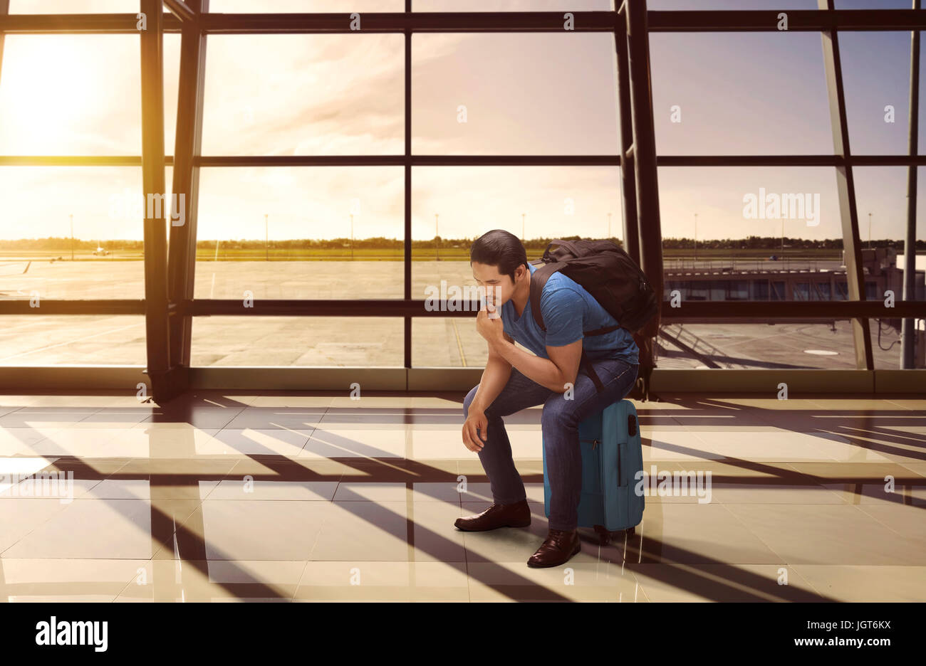 Confused asian tourist sitting on suitcase waiting departure in the airport Stock Photo