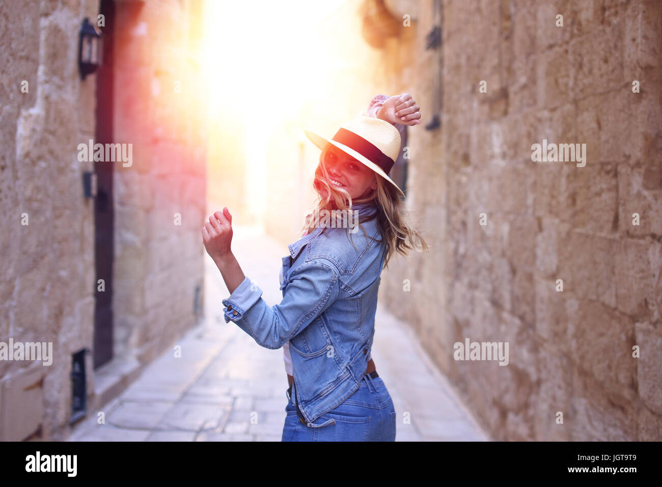 Young woman in jeans and hat dancing in sunset on old narrow streeet Stock Photo