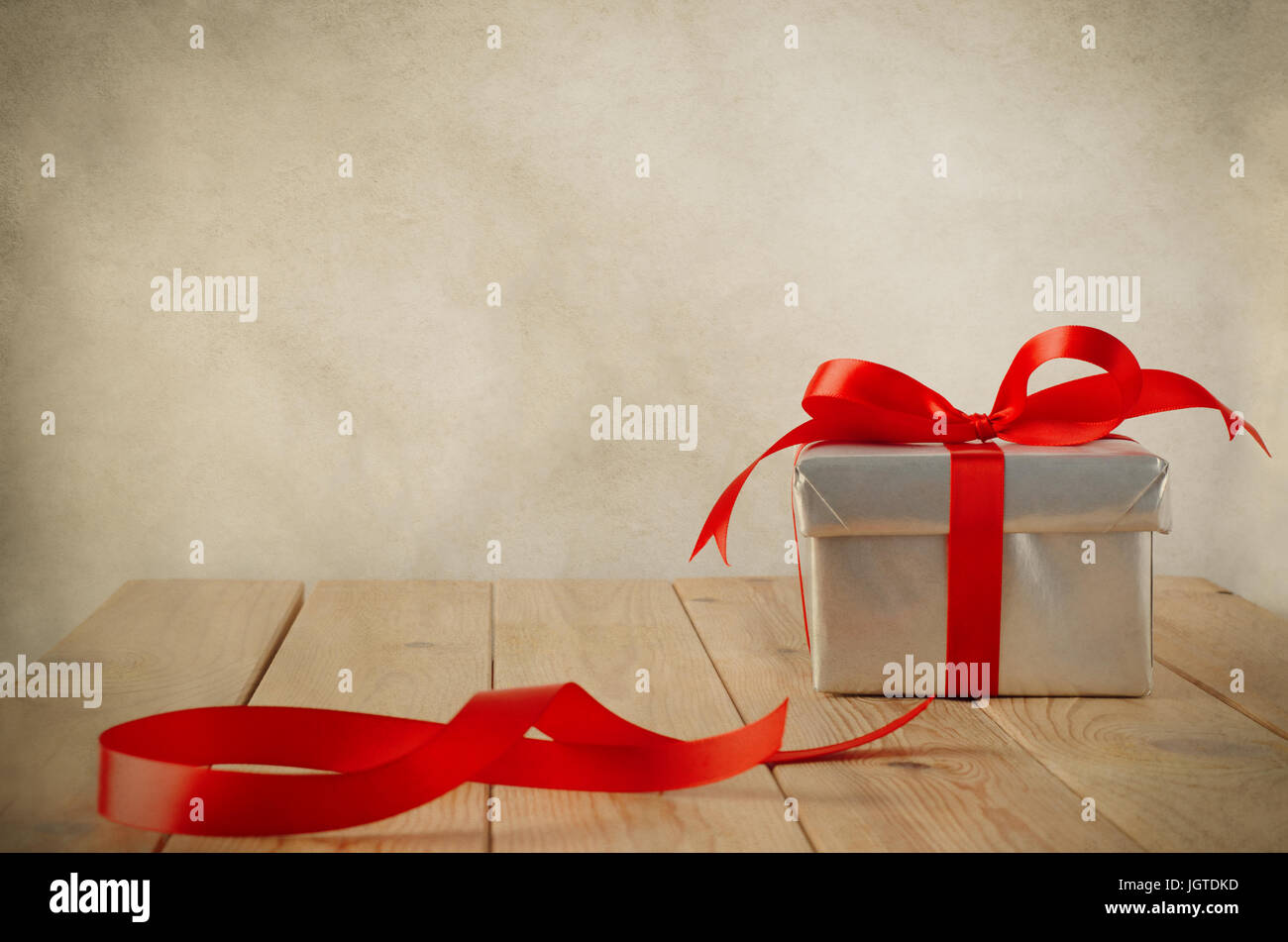 A Christmas gift box with closed lid, wrapped in silver paper and tied to a bow with a red satin ribbon.  Placed on a weathered old wooden table with  Stock Photo