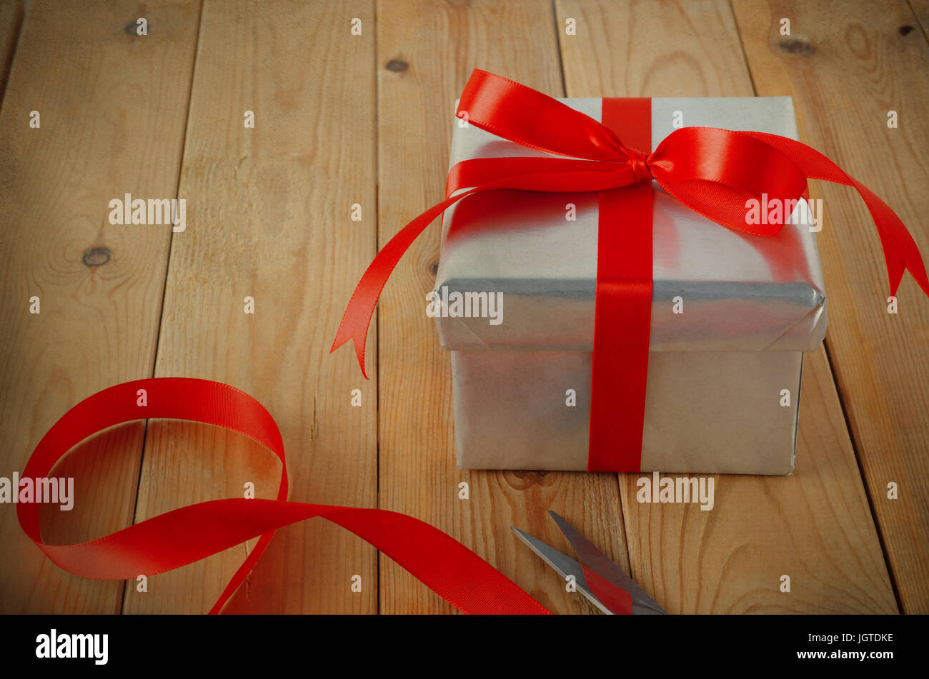 Gift wrapping scene. A Christmas gift box wrapped in silver paper and tied with red ribbon to a bow, on an old wood plank table with scissors and ribb Stock Photo