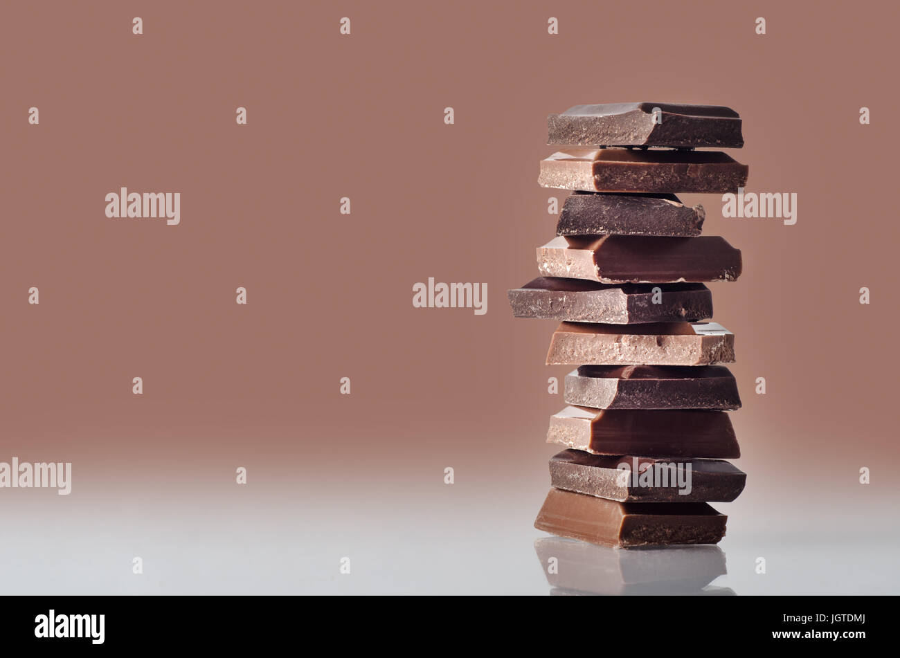 Eye level shot of chocolate pieces, broken from a bar and stacked in a column, standing on a reflective surface against a chocolate brown background w Stock Photo