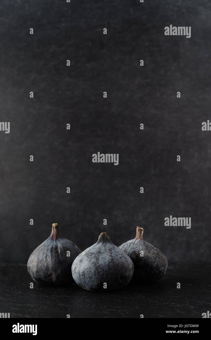 Three figs grouped in a still life composition on black slate surface with dusty black background.  Shot just above eye level. Stock Photo
