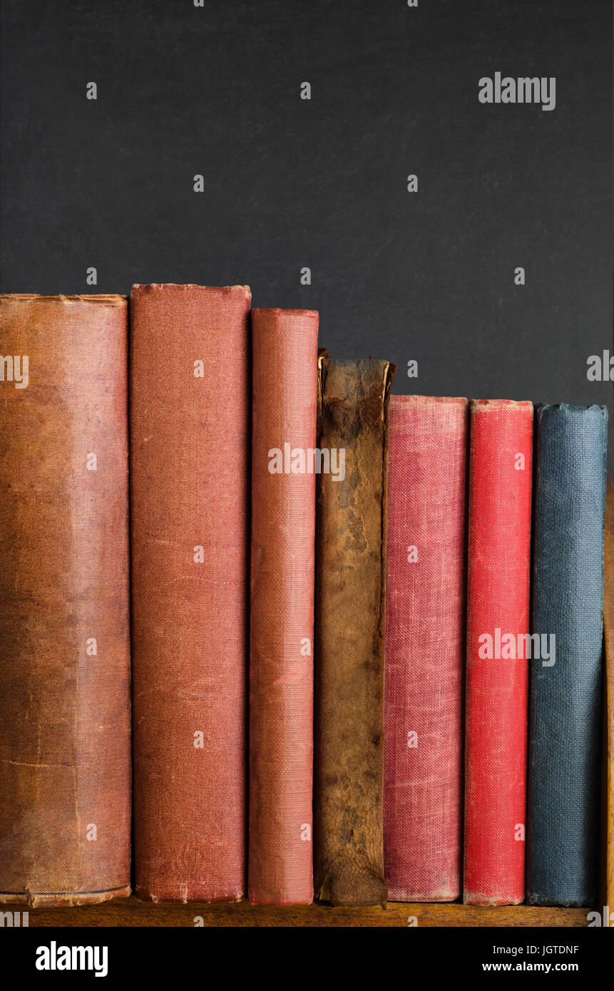 Wooden bookshelf with row of battered old vintage books standing upright.  Blank spines and chalkboard background provide copy space. Stock Photo