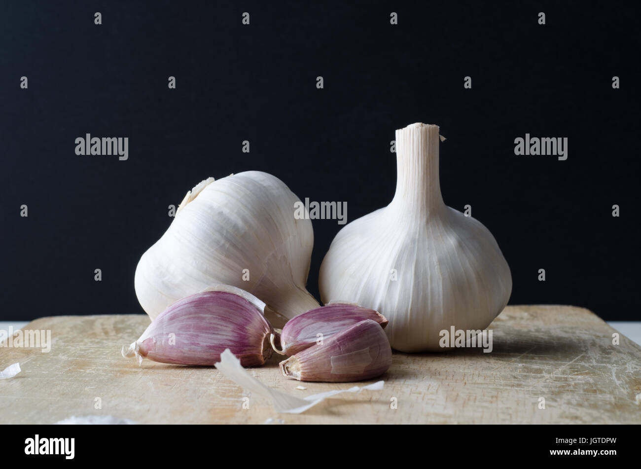 Two whole, unpeeled garlic bulbs and three pink garlic cloves on a scratched old wooden chopping board, with outer peelings scattered around.  Black b Stock Photo
