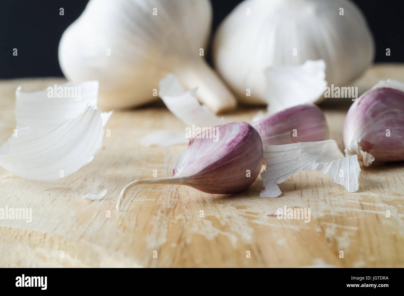 Two whole garlic bulbs in the background, with  pink cloves in foreground on a wooden chopping board surrounded by scattered fragments of shedded pape Stock Photo