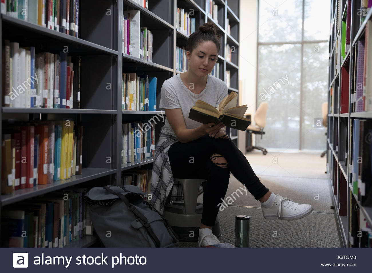 Female college student reading book among bookshelves in library Stock Photo Alamy