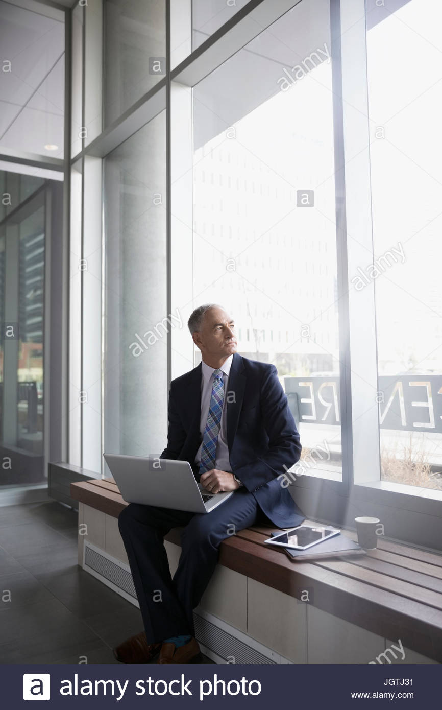 Pensive Businessman Using Laptop On Bench In Modern Office Lobby