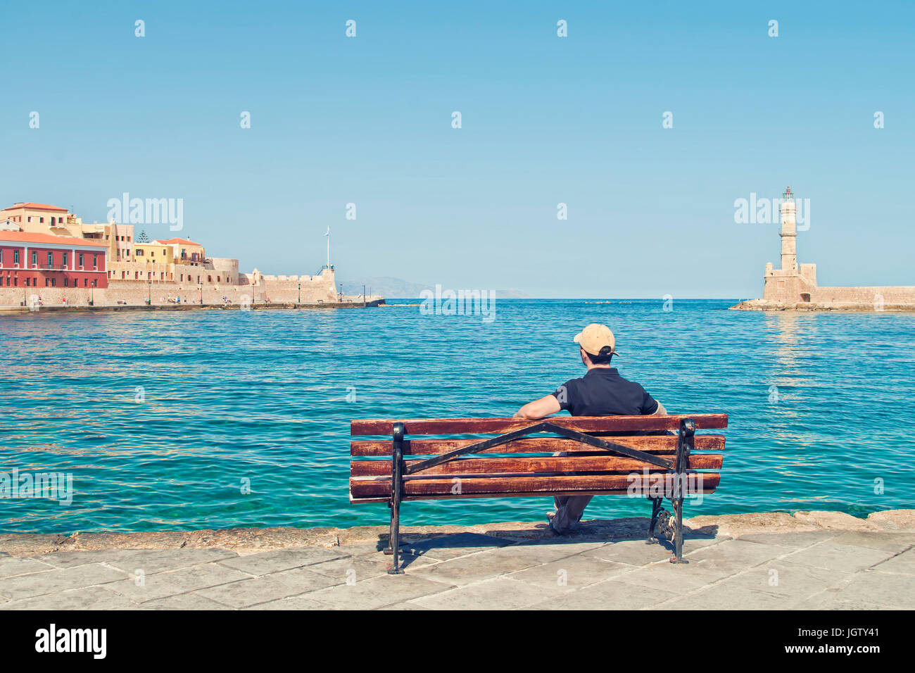 young caucasian male tourist wearing black t-shirt and baseball cap sitting alone on bench and looking at old part of Chania and venetian lighthouse o Stock Photo