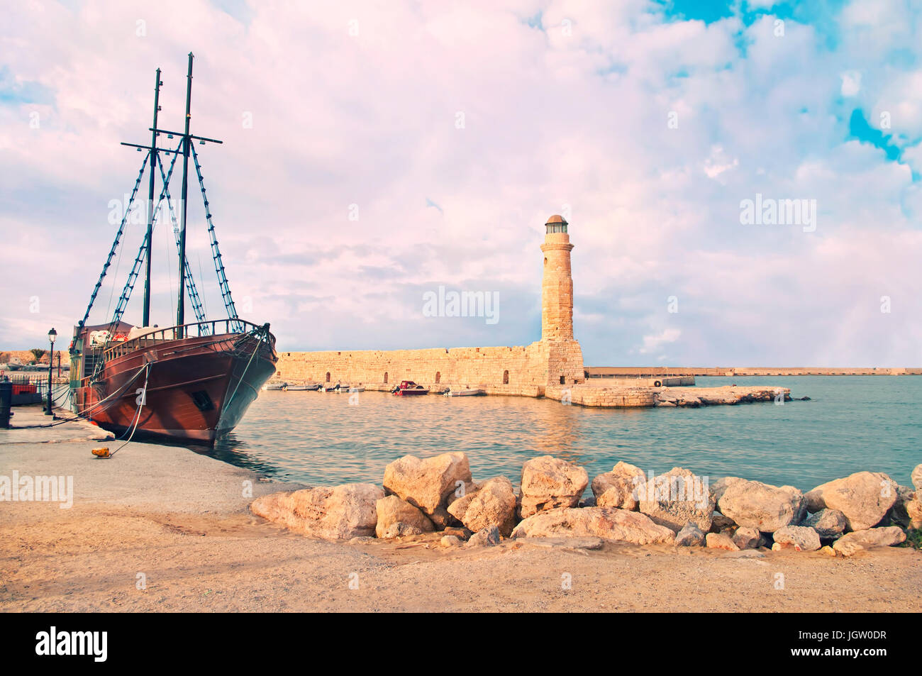 cross-processed image of old Venetian Lighthouse with old-style wooden ship moored in dock in warm sunset colours, Rethymno, Crete, Greece Stock Photo