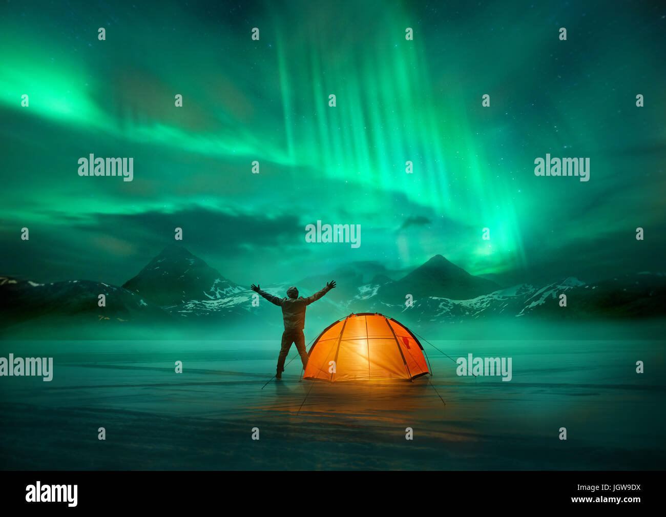 A man camping in wild northern mountains with an illuminated tent viewing a spectacular green northern lights aurora display. Photo composition. Stock Photo