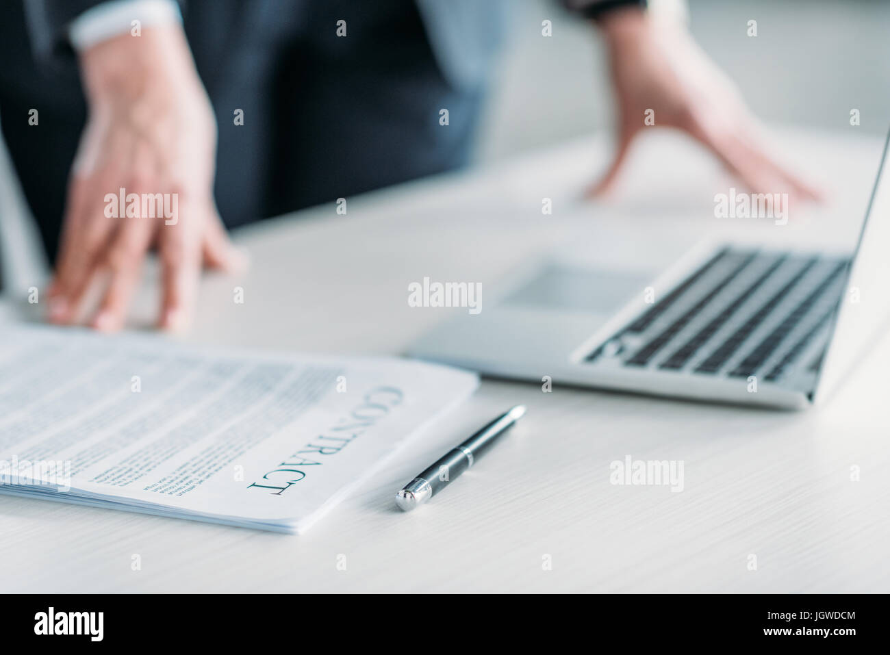 laptop computer, pen and contract on tabletop, business person behind