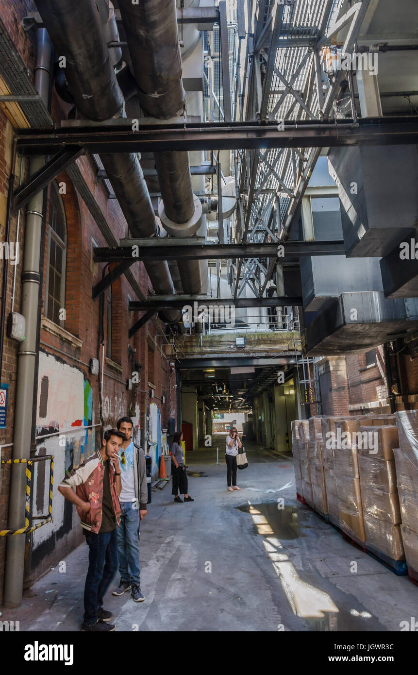 Looking along an open corridor which contains people smoking, puddles, piles of bricks, metal ducting, pipes and a metal walkway. Stock Photo