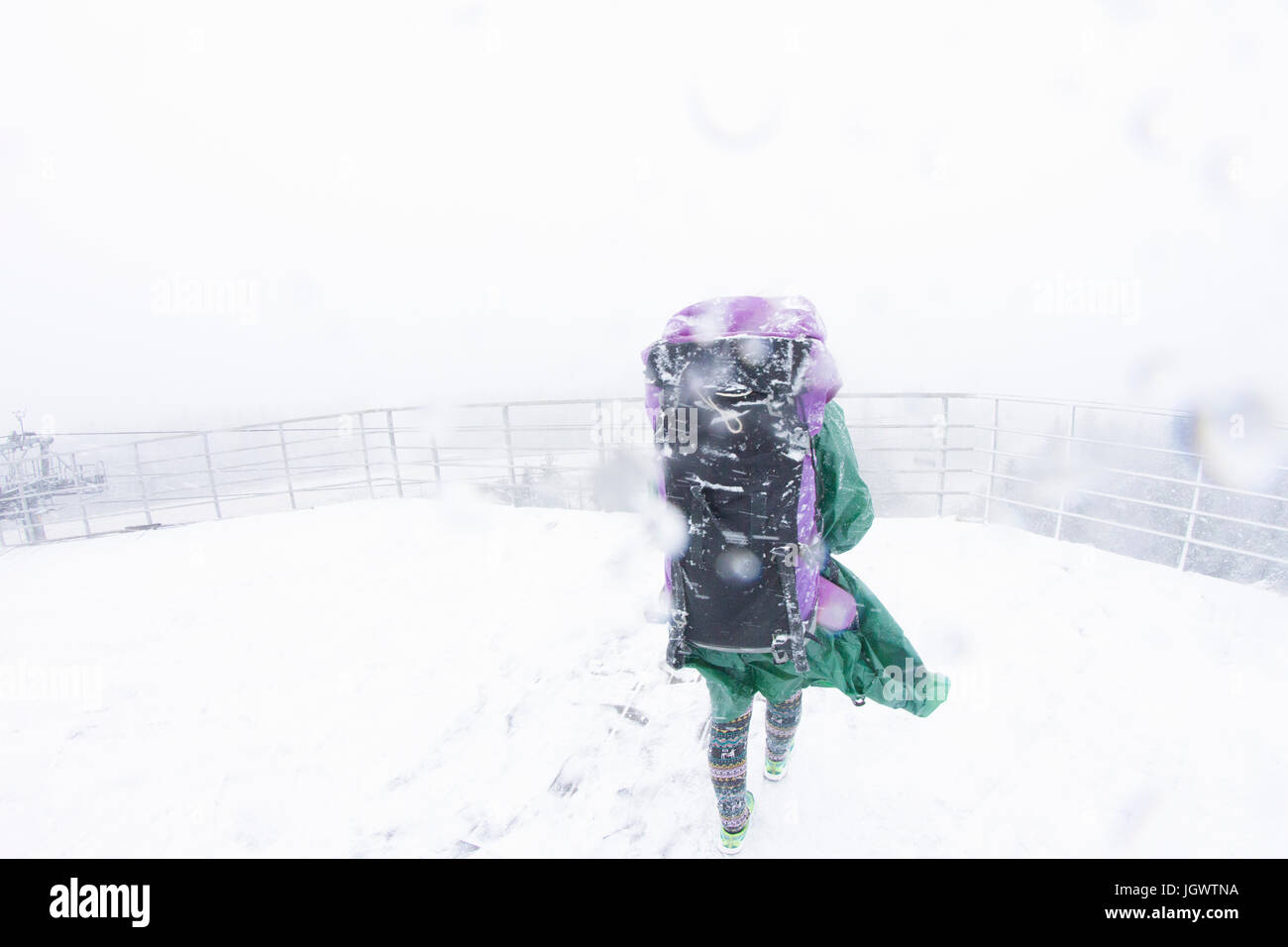 Rear view of female hiker in waterproof clothing walking on mountain viewing platform in snowstorm Stock Photo