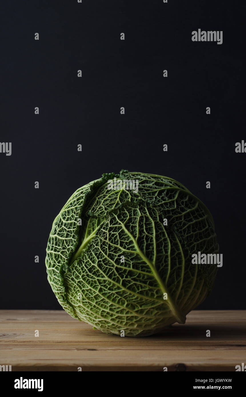 A whole green cabbage on a raw wood planked table, against a black chalkboard background.  Moody lighting. Stock Photo