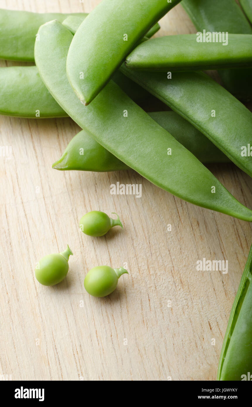 A pile of pea pods, with three tiny baby peas extracted and placed on a light wooden table.  Stalks intact. Stock Photo