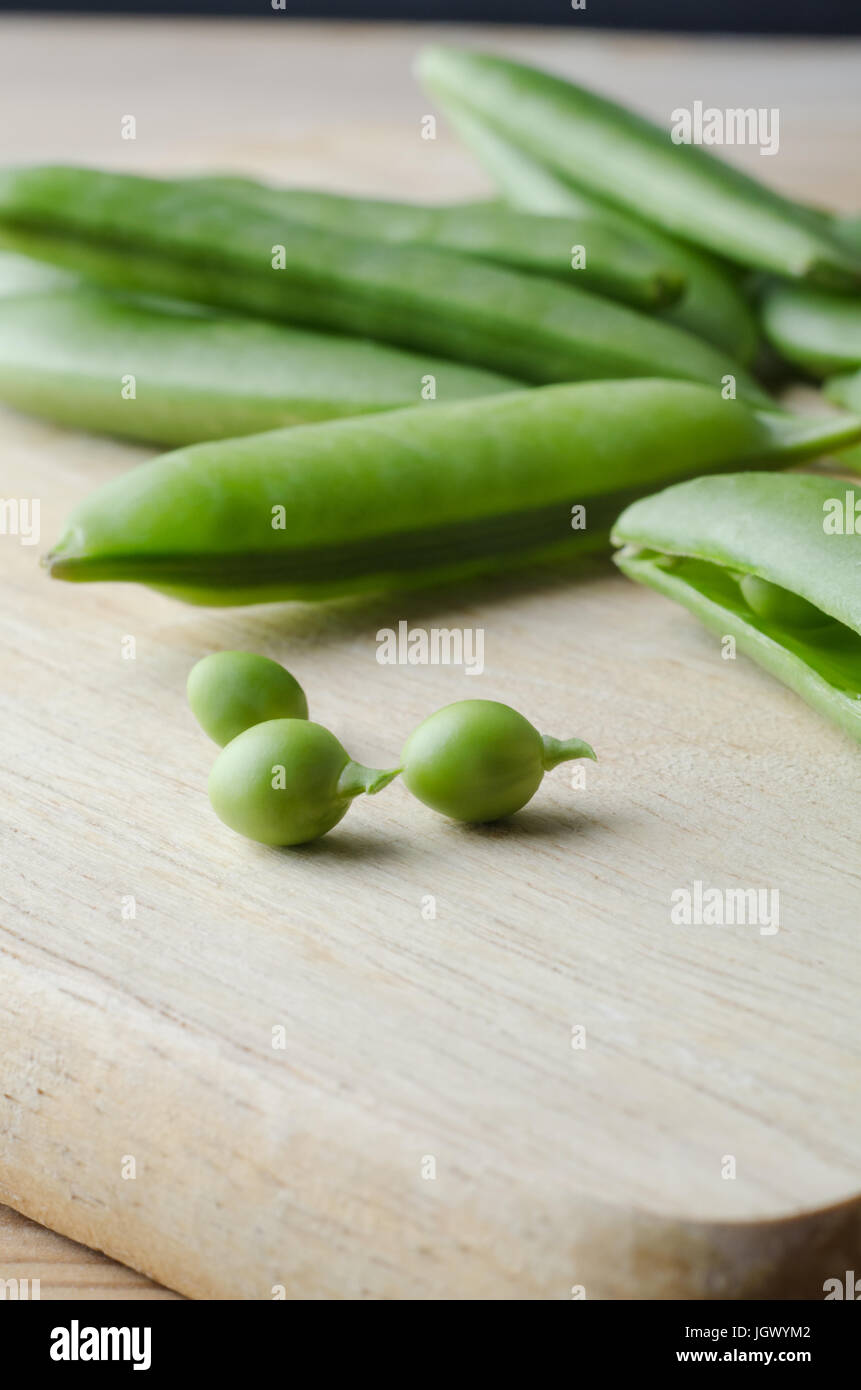 Close up of fresh, raw pea pods, scattered on a wooden chopping board, with three small peas extracted and placed in the foreground with stalks intact Stock Photo